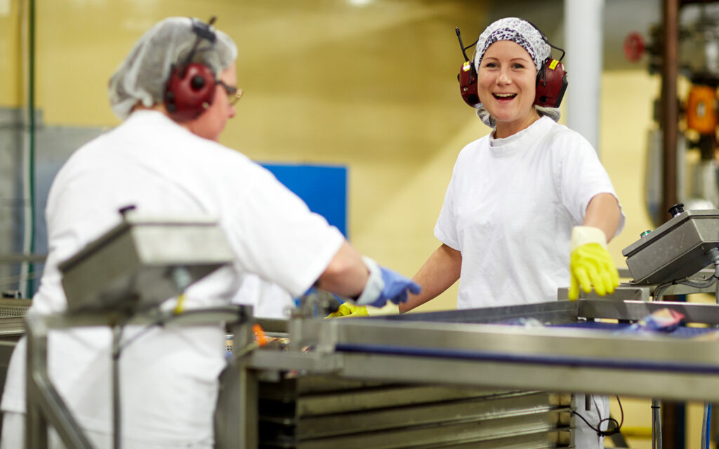 Två medarbetare som står vid ett produktionsband. Vitklädda, med hörselkåpor, hårnät och handskar.