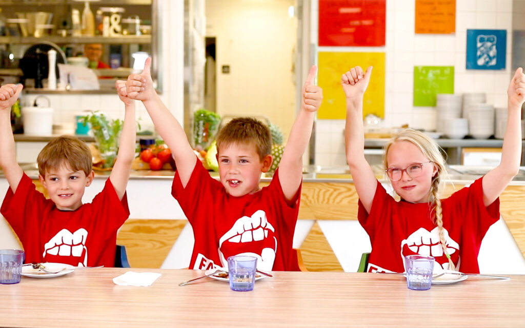 Tre barn med Fast Fusion Friday-tshirts sitter vid ett bord och ger tummarna upp till den goda maten de just ätit.