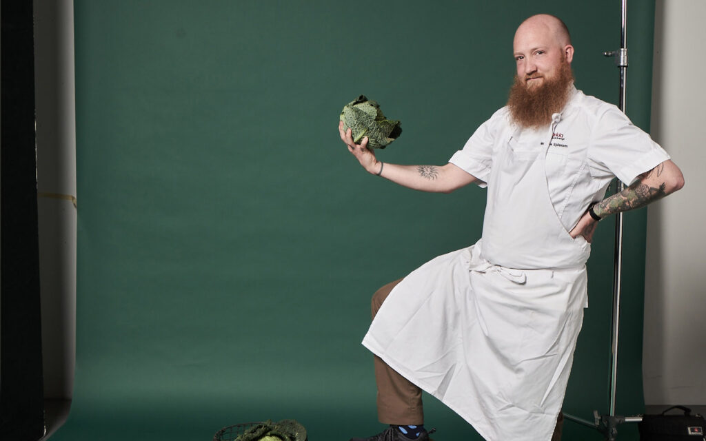 Kocken Mattias Sjöblom poserar med en fot på en låda och ett blomkålshuvud i handen.