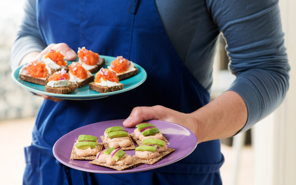 Människa i blått förkläde som håller två tallrika med näringsrika mellanmål. Recept från konceptet Snackisar.