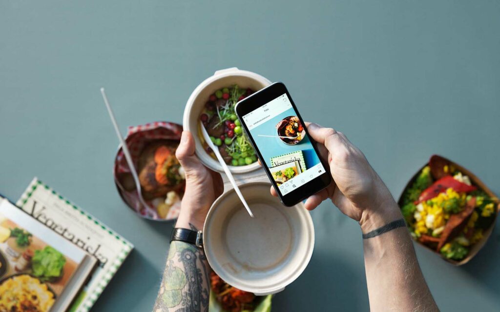 Olika maträtter mot en grå-blå bakgrund. Hand håller i bowl och i en mobil samtidigt.
