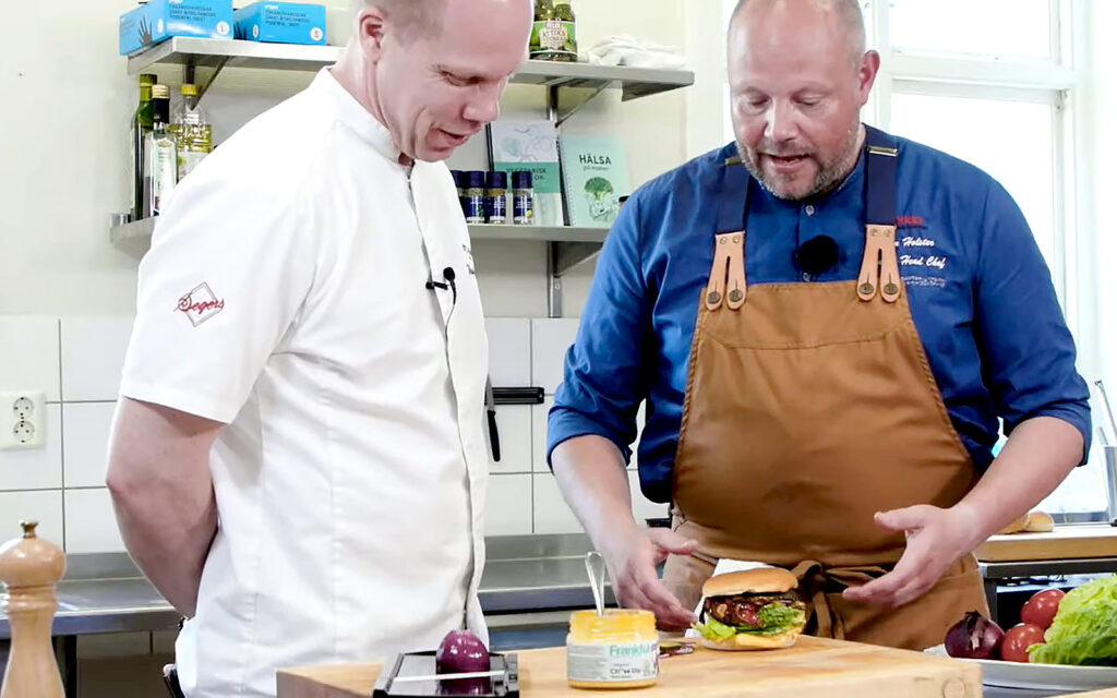 Kockarna Pim och Johan Holster står vid en bänk. Johan greppar efter en burgare. Bredvid står en burk med Frankful Ch**se Dip.