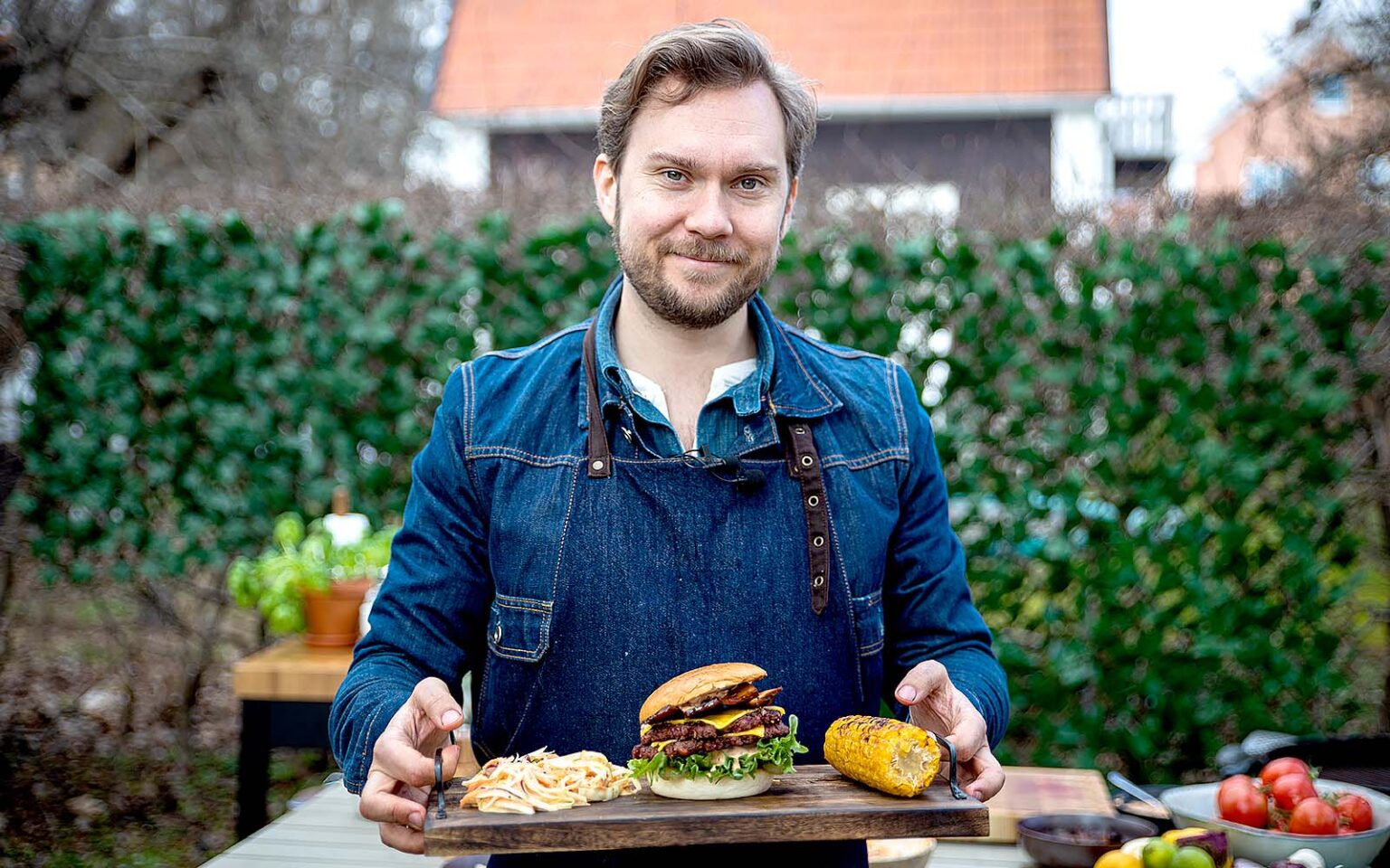 Gustav "Jävligt Gott" Johansson står utomhus i en trädgård. Han håller en bricka med en burgare, cole slaw och en majskolv på.