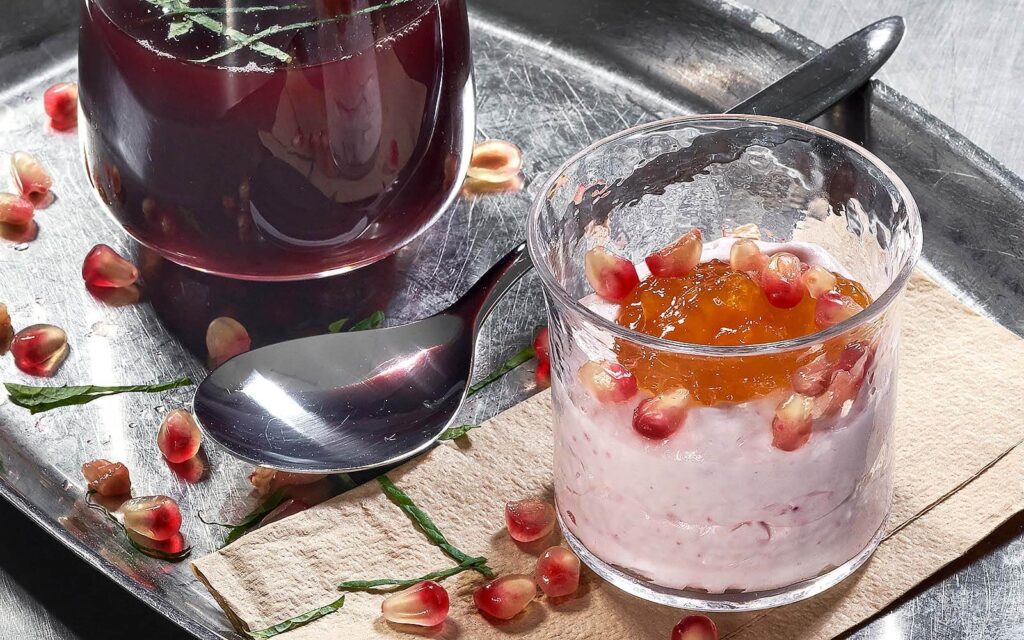 Glas med Ekströms Mousse och Kompott, garnerad med granatäppelkärnor. I bakgrunden syns en sked och ett glas med röd dryck.