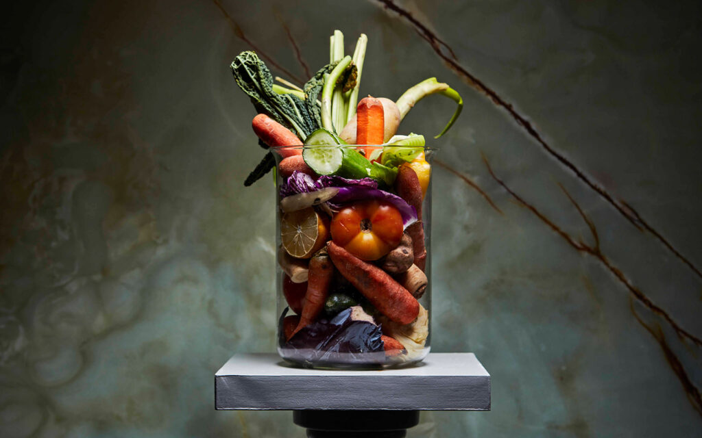 Cylindervas i glas på en piedestal, vasen är full med gamla grönsaker.