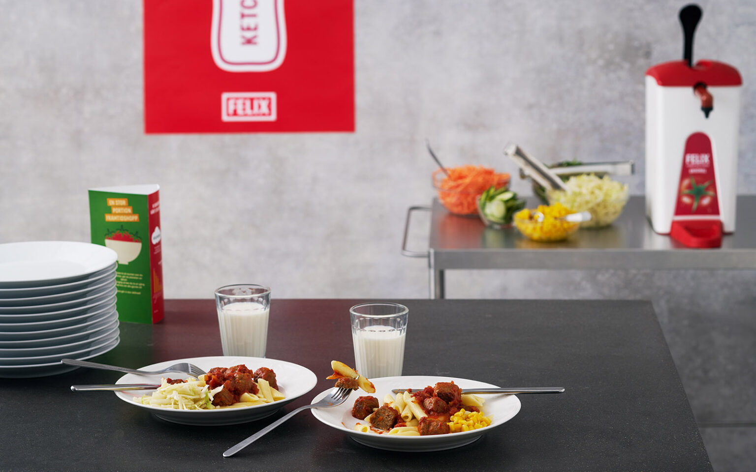 Två tallrikar med FELIX Nötköttbulle med ärta med pasta och FELIX Tomatketchup. I bakgrunden syns staplar med tallrikar, en serveringsvagn med sallad, en Sentomat med FELIX Tomatketchup och en affisch från FELIX.