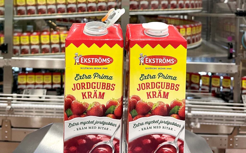 Två förpackningar med Ekströms Extra Prima Jordgubbskräm i en fabrik där det syns mängder av liknande förpackningar i bakgrunden.