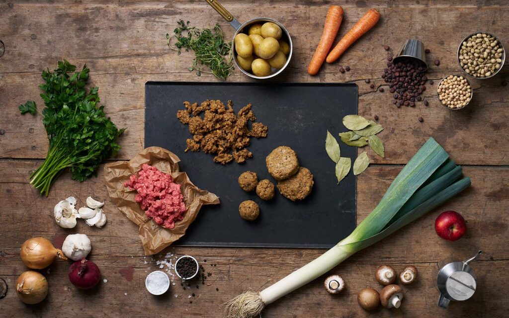 Växtbaserad färs och en massa olika ingredienser, råvaror och grönsaker ligger på ett träbord.