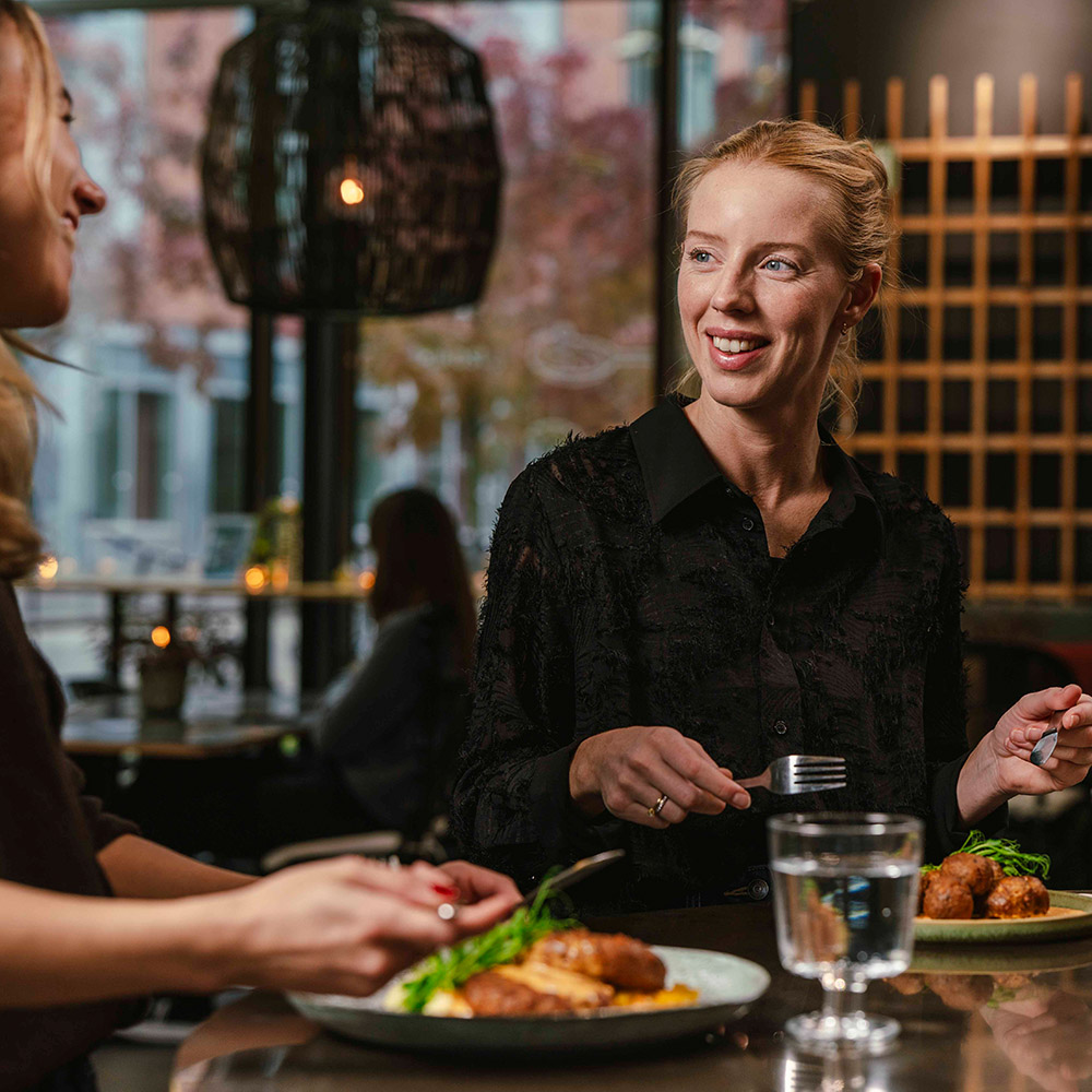 Två kvinnor sitter i en restaurang och äter 50/50-rätter och ler mot varandra.