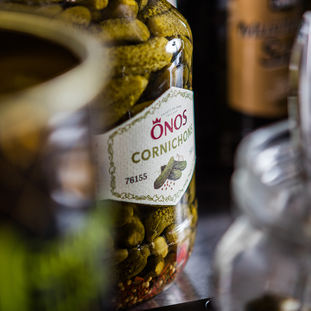 Burk med Önos Cornichons på en bänk tillsammans med andra produkter som är ur fokus.