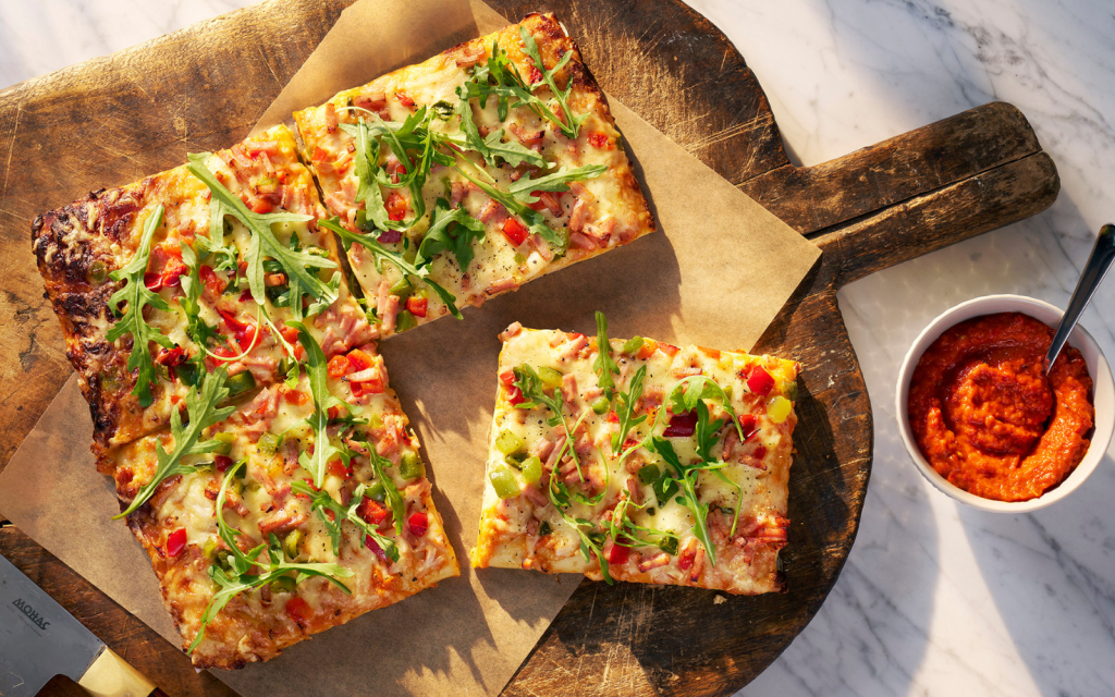 Fyrkantspizza Skinka på en träbricka med tillbehör som rucola och ajvar